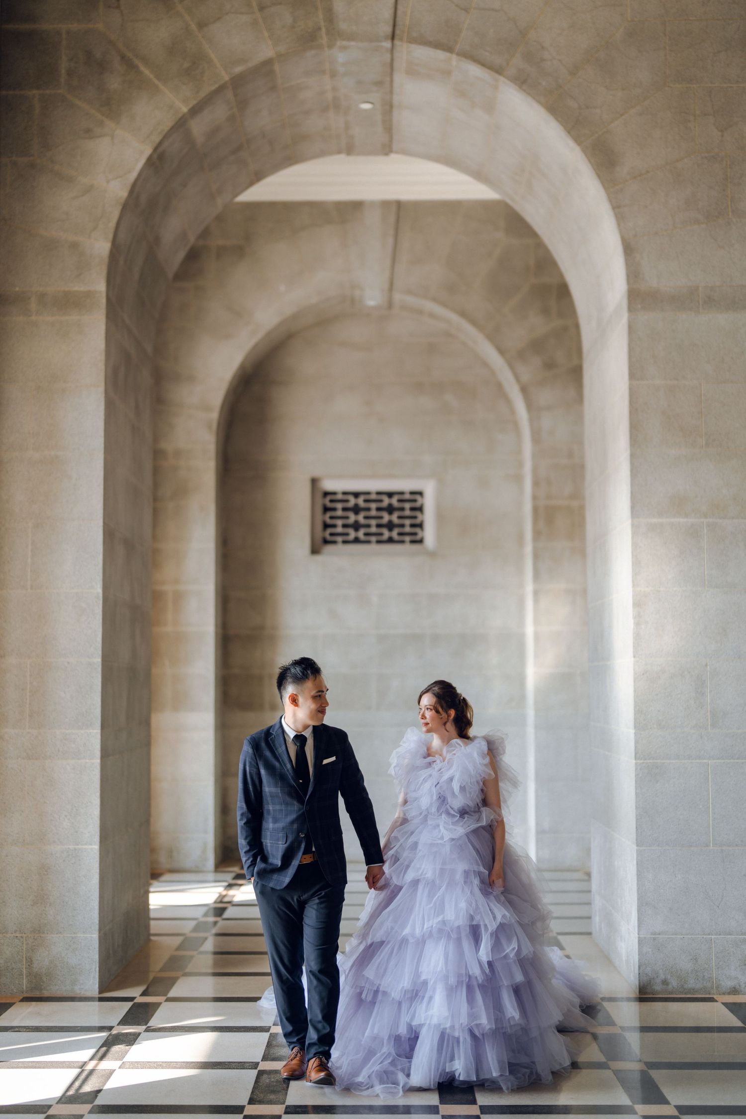 A couple in wedding attire stands in an elegant stone archway with dramatic lighting and shadows.