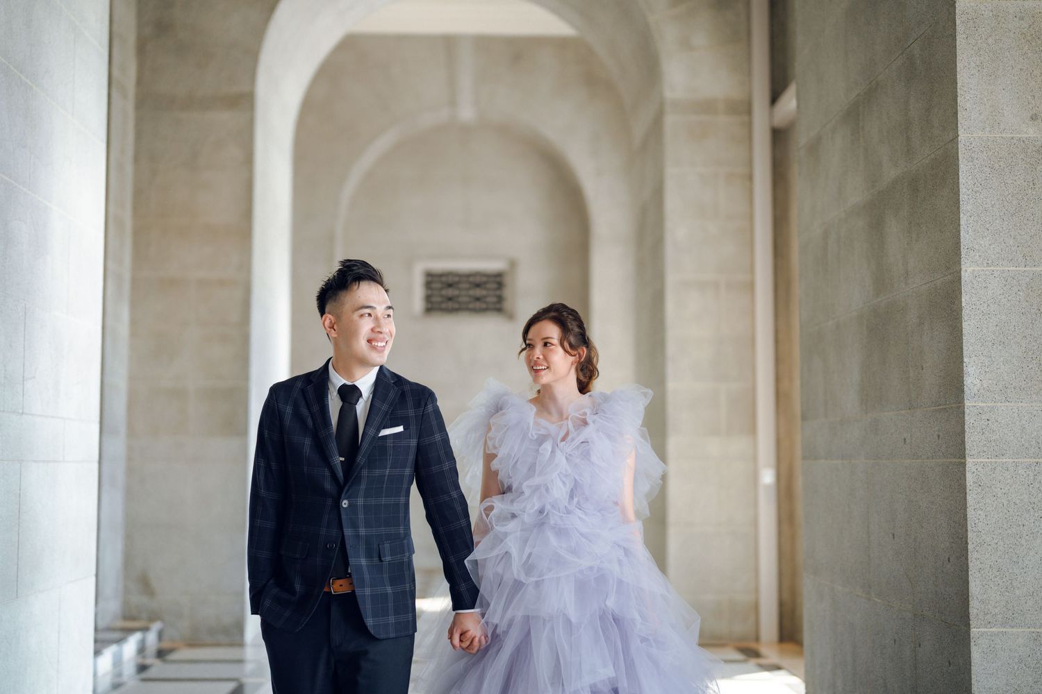 Wedding couple walks hand in hand through a grand stone colonnade hallway.