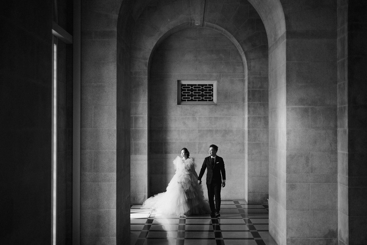 Black and white wedding photo showing a couple walking through an elegant stone hallway.