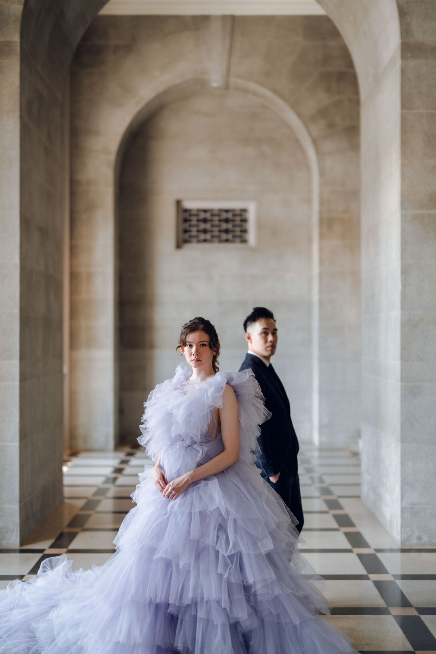 Elegant couple poses in formal attire against a grand colonnade with arched stone architecture.