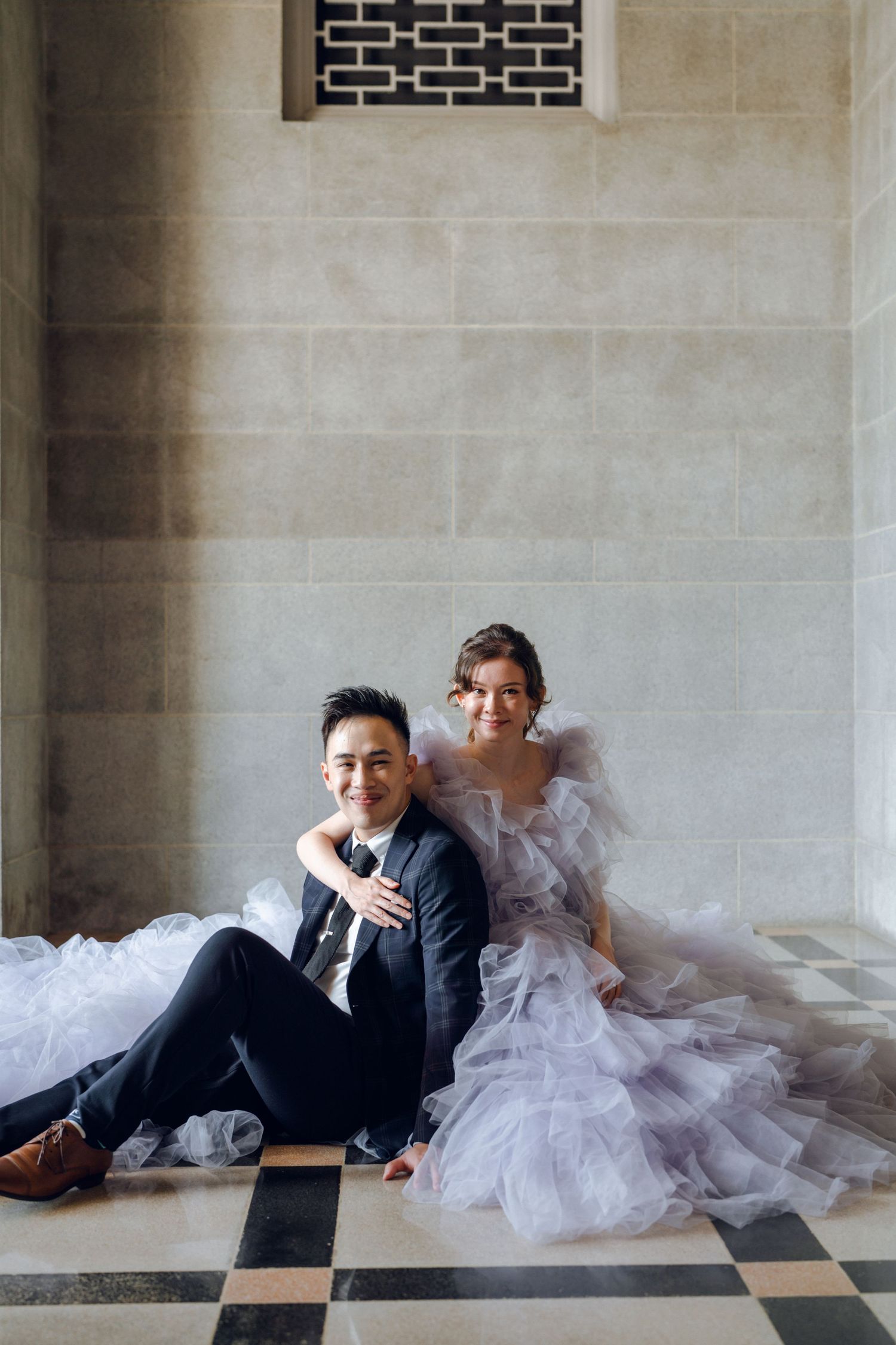 Couple relaxes on checkered floor against stone wall in formal wedding attire.
