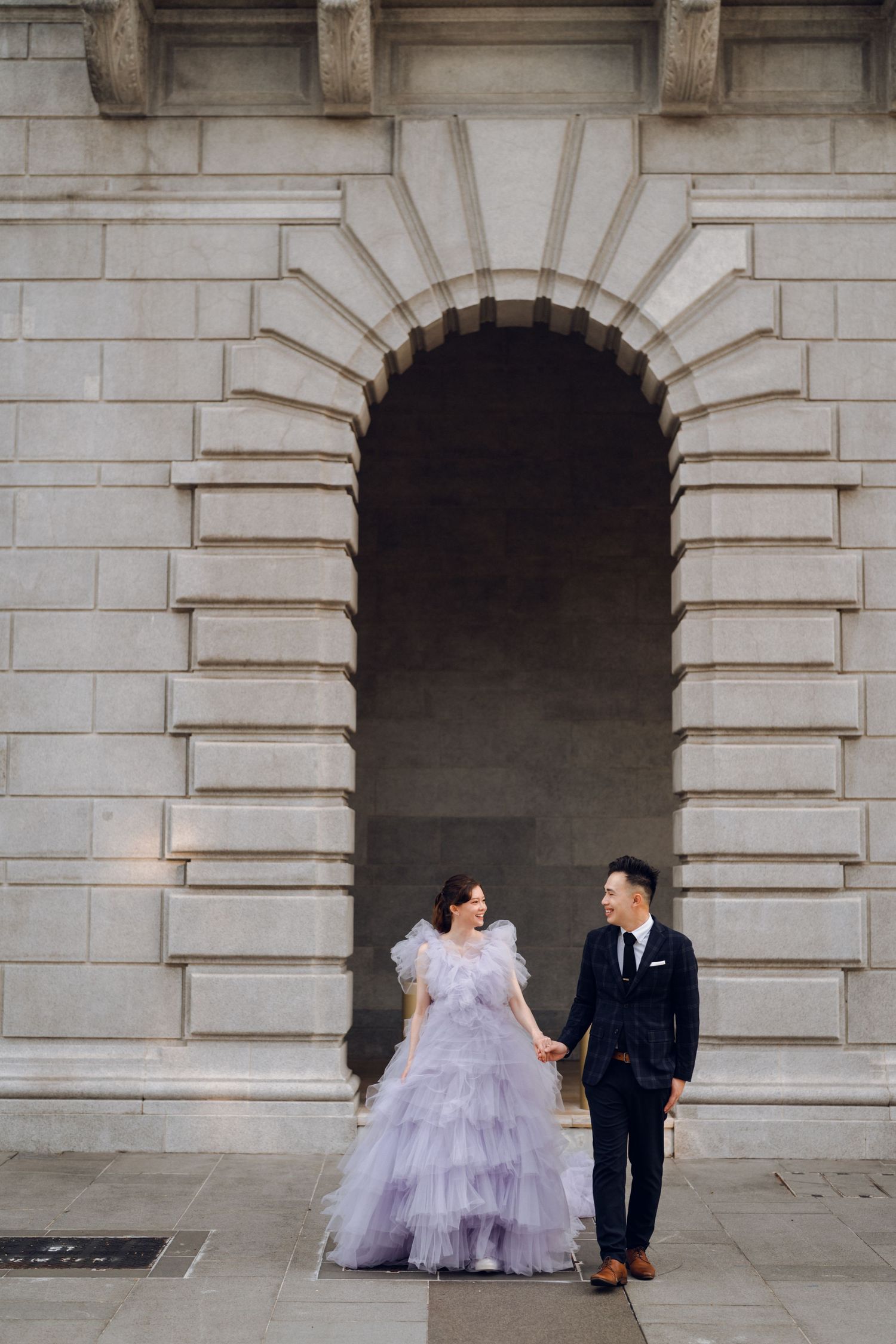 A couple stands beneath an ornate stone archway in formal wedding attire.