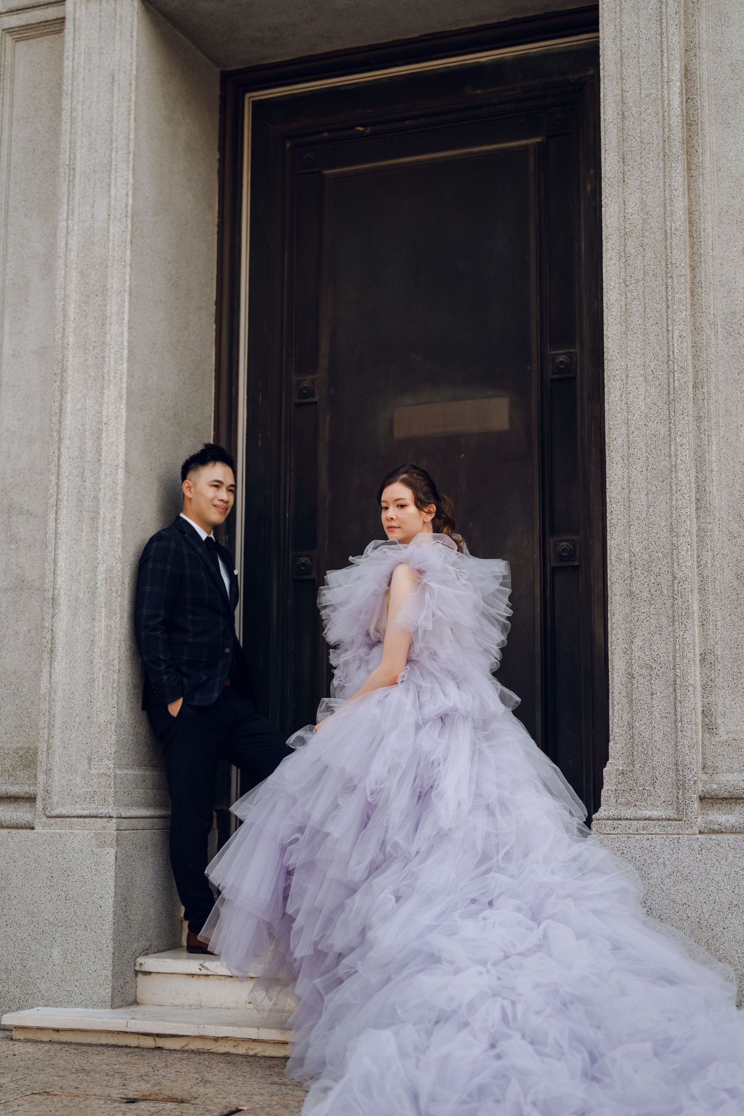 A lavender tulle ball gown flows dramatically against classical stone columns in an elegant architectural setting.