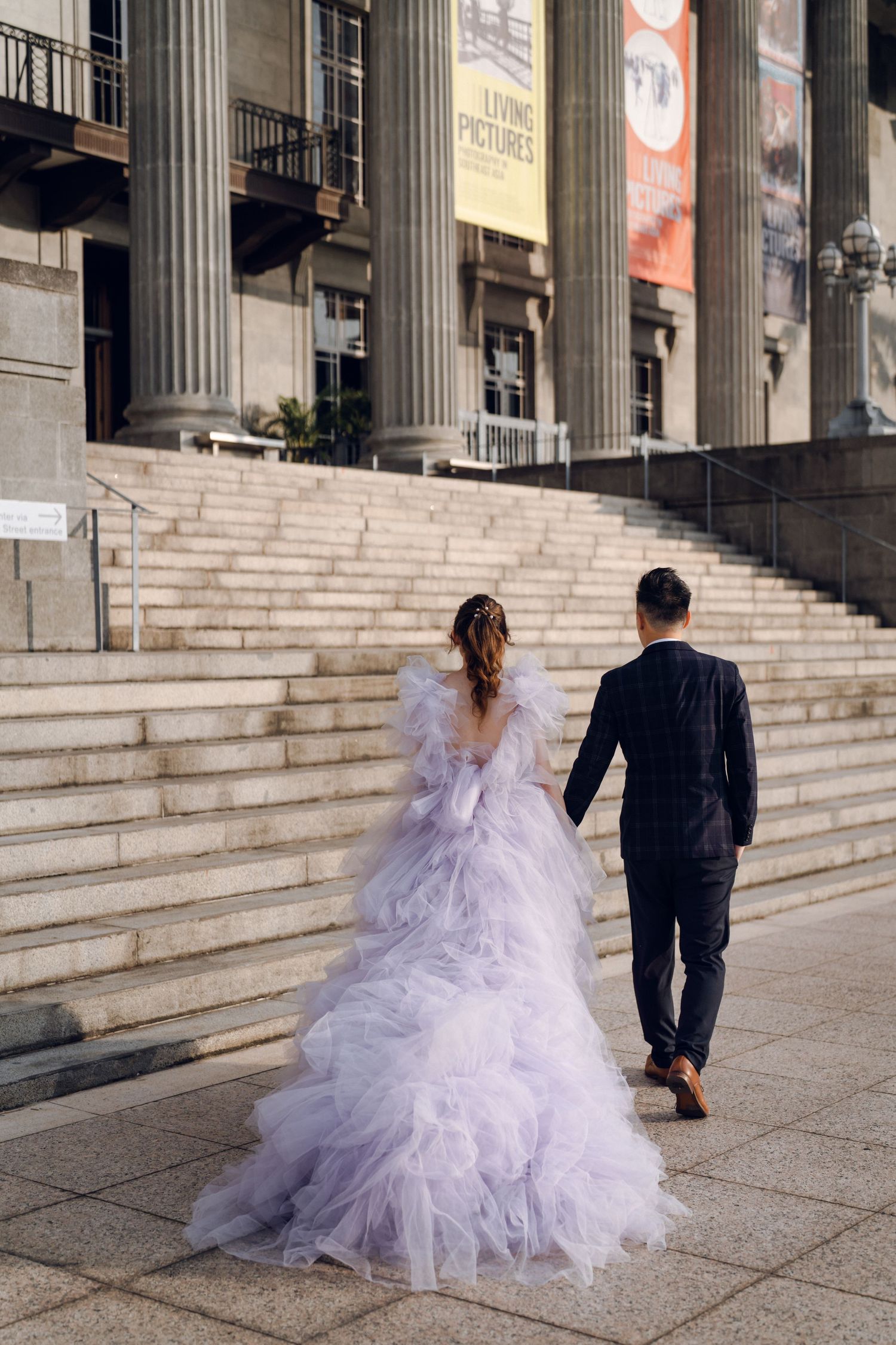 A couple walks up grand stone steps, with a dramatic lavender gown trailing behind.
