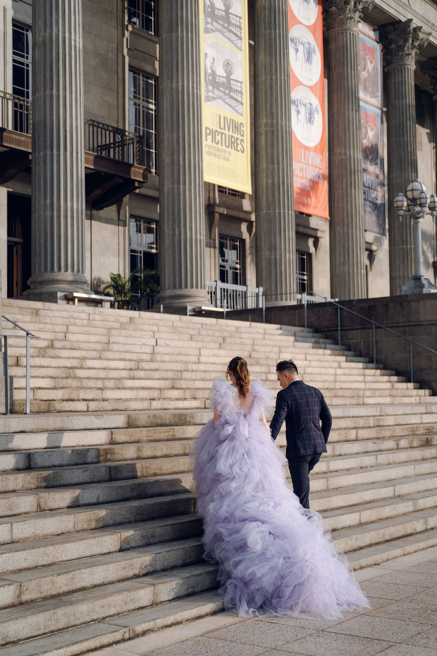 Two figures ascend majestic stone steps of a classical building, one wearing a flowing lavender gown.