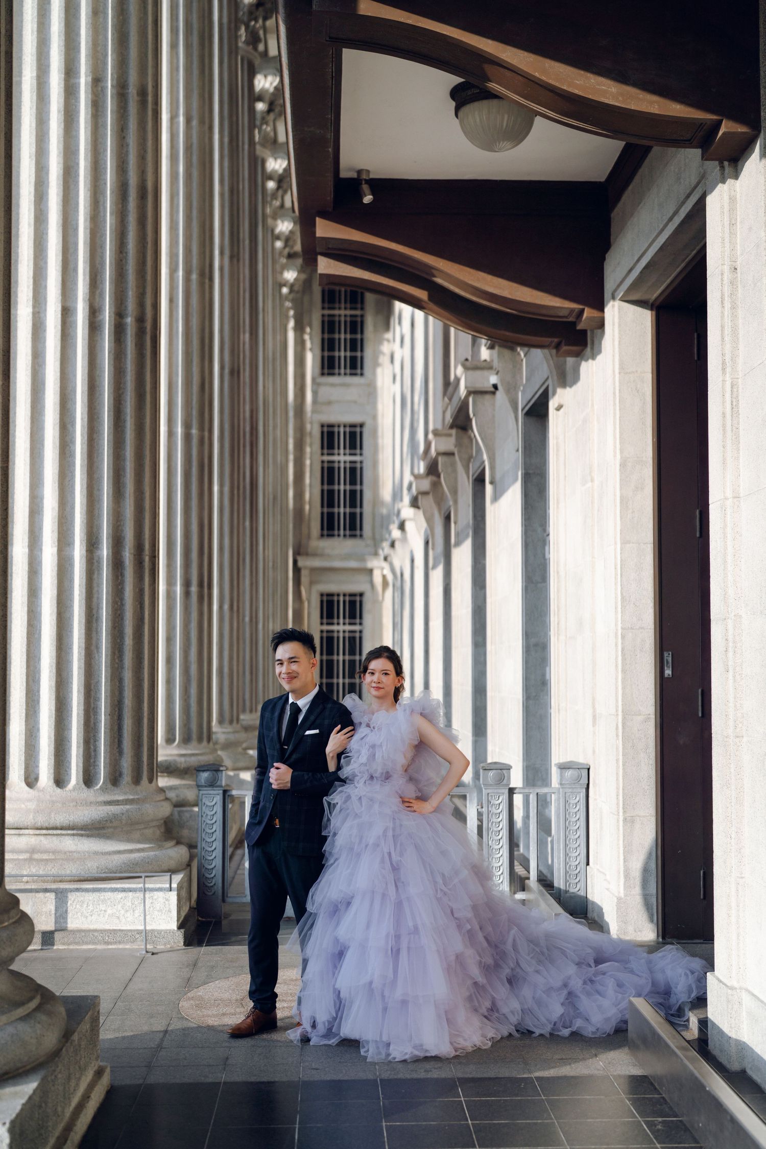 An elegant couple poses between classical stone columns, with a flowing lavender gown as centerpiece.