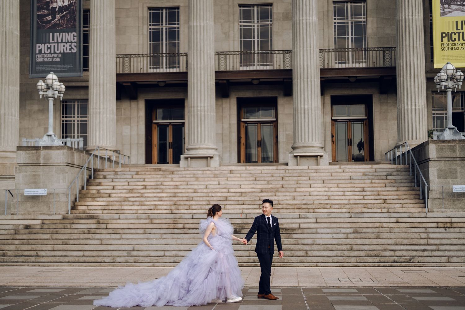 A couple poses on grand stone steps outside a historic building, with the bride wearing a voluminous lavender gown.