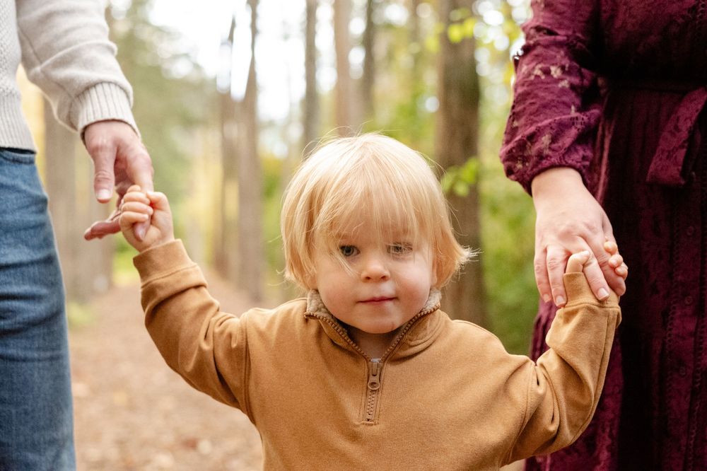 A small child in a cream sweater walks through autumn woods while holding hands with adults on both sides.