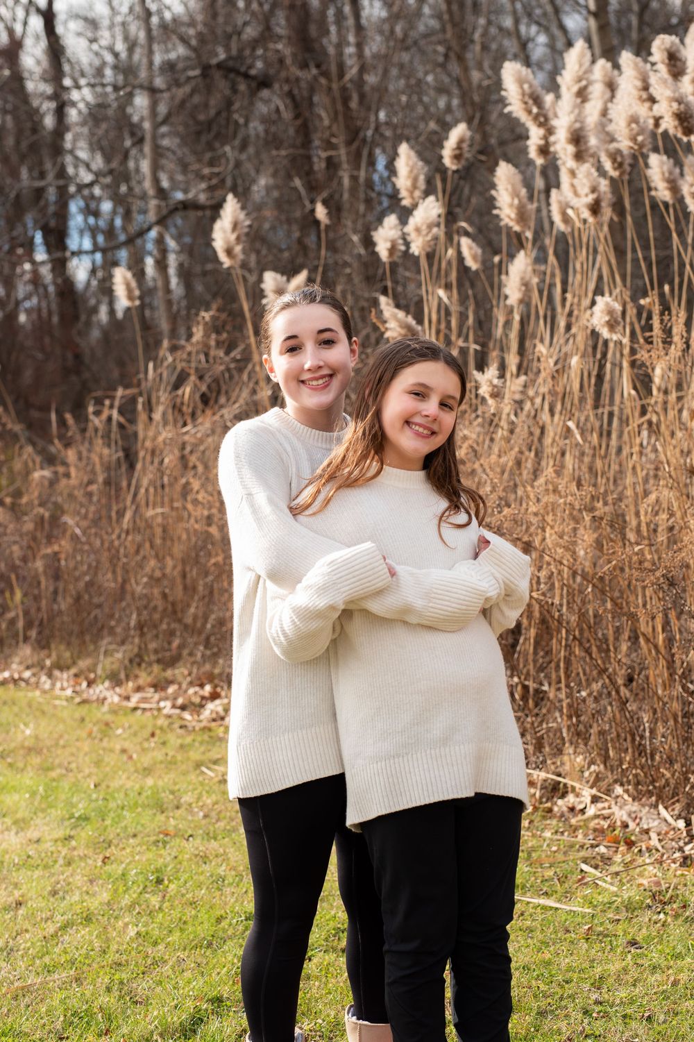 Two people in matching cream sweaters and black pants embrace while standing in tall autumn grass.