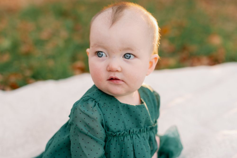 A baby in a dark green dress sitting on a white blanket during fall.