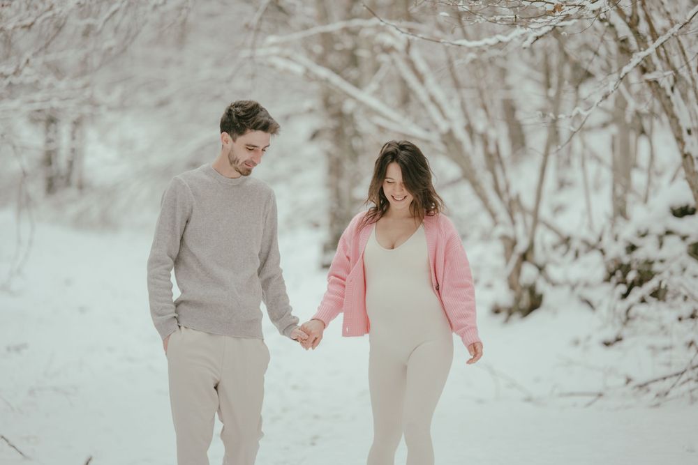 Couple holding hands while walking through a snow-covered forest path in complementary winter outfits.