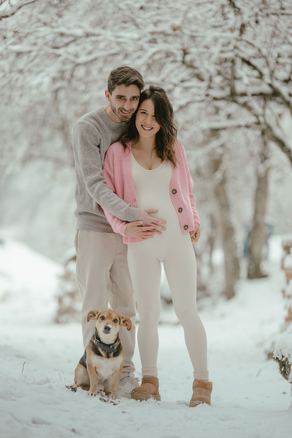 A couple poses with their dog in a snow-covered landscape wearing cozy winter sweaters.