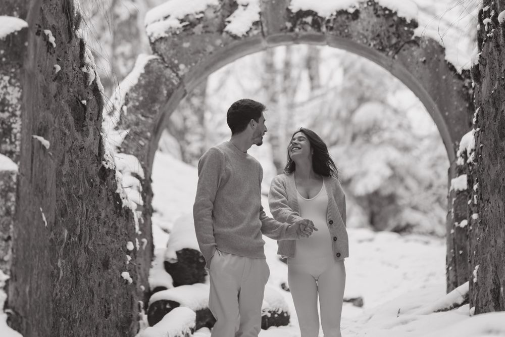 A couple stands beneath a snow-covered stone archway in a winter landscape.