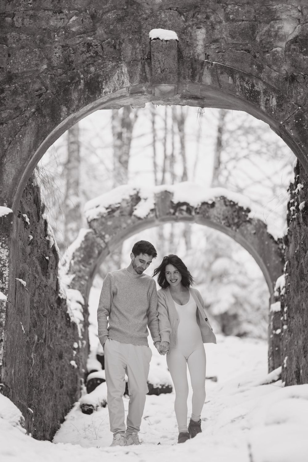 A couple walks through connected stone archways in a snowy winter setting.