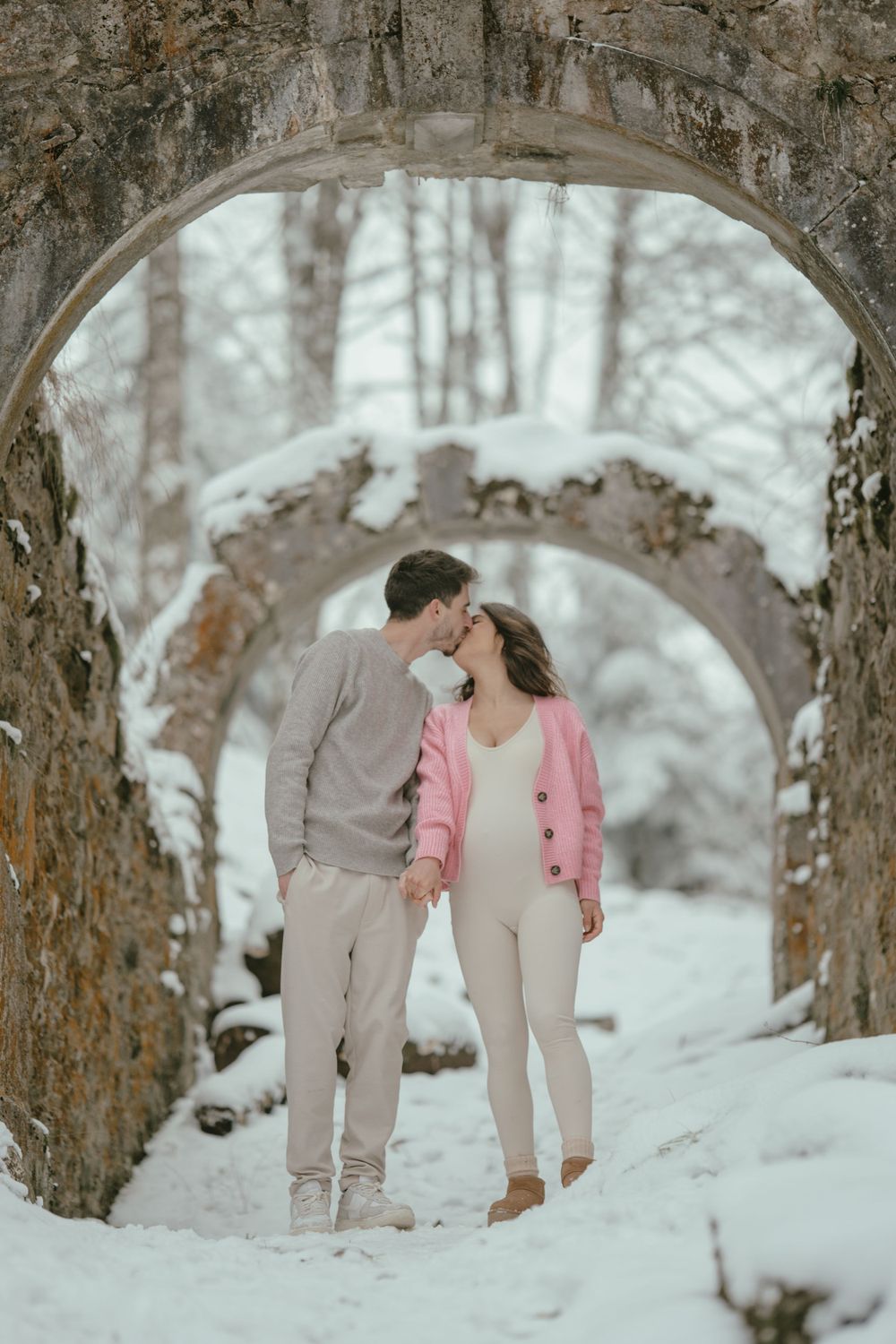 A couple shares a kiss under stone archways surrounded by snow-covered trees.