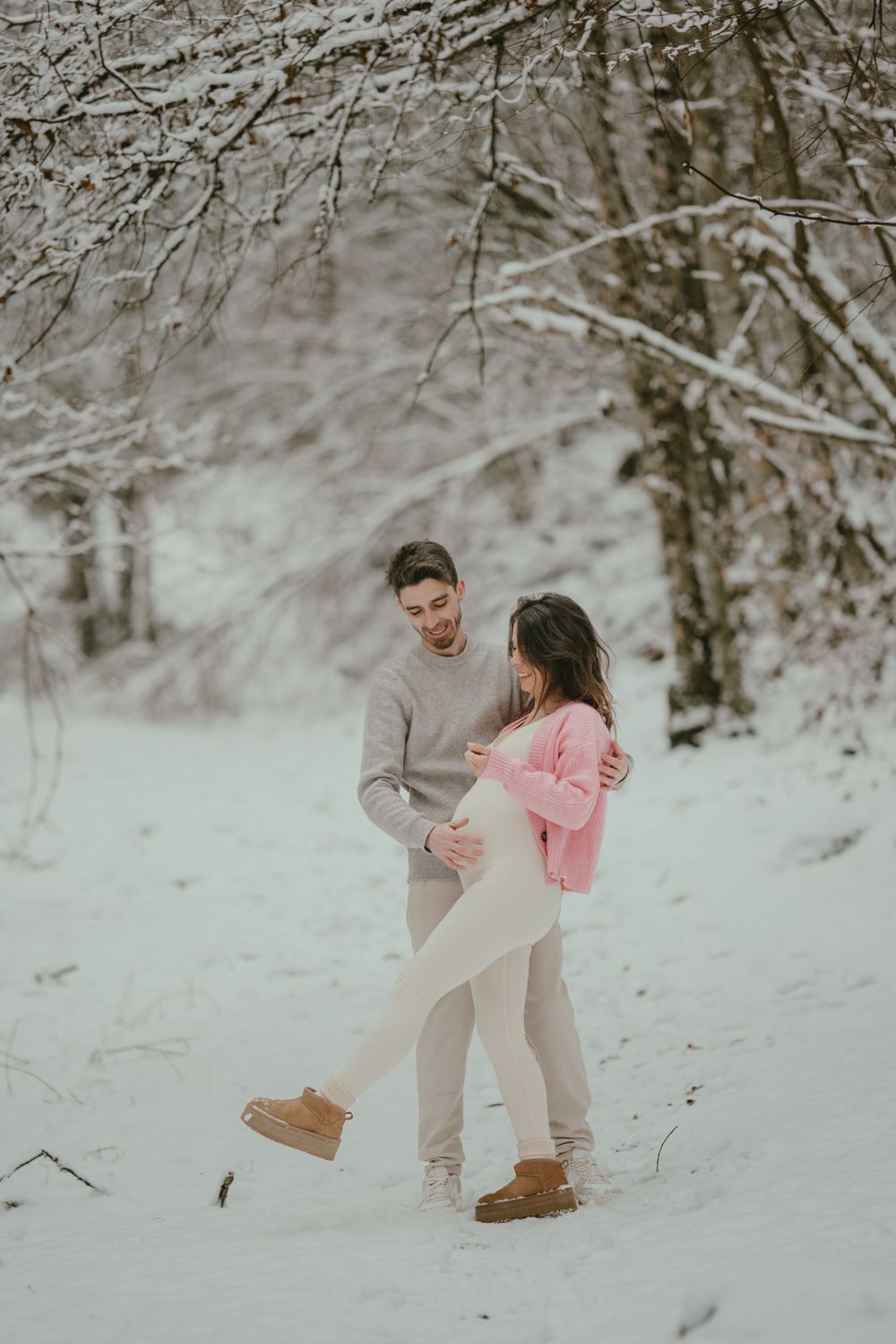 A couple embraces in a snowy winter forest setting wearing coordinated beige and pink outfits.