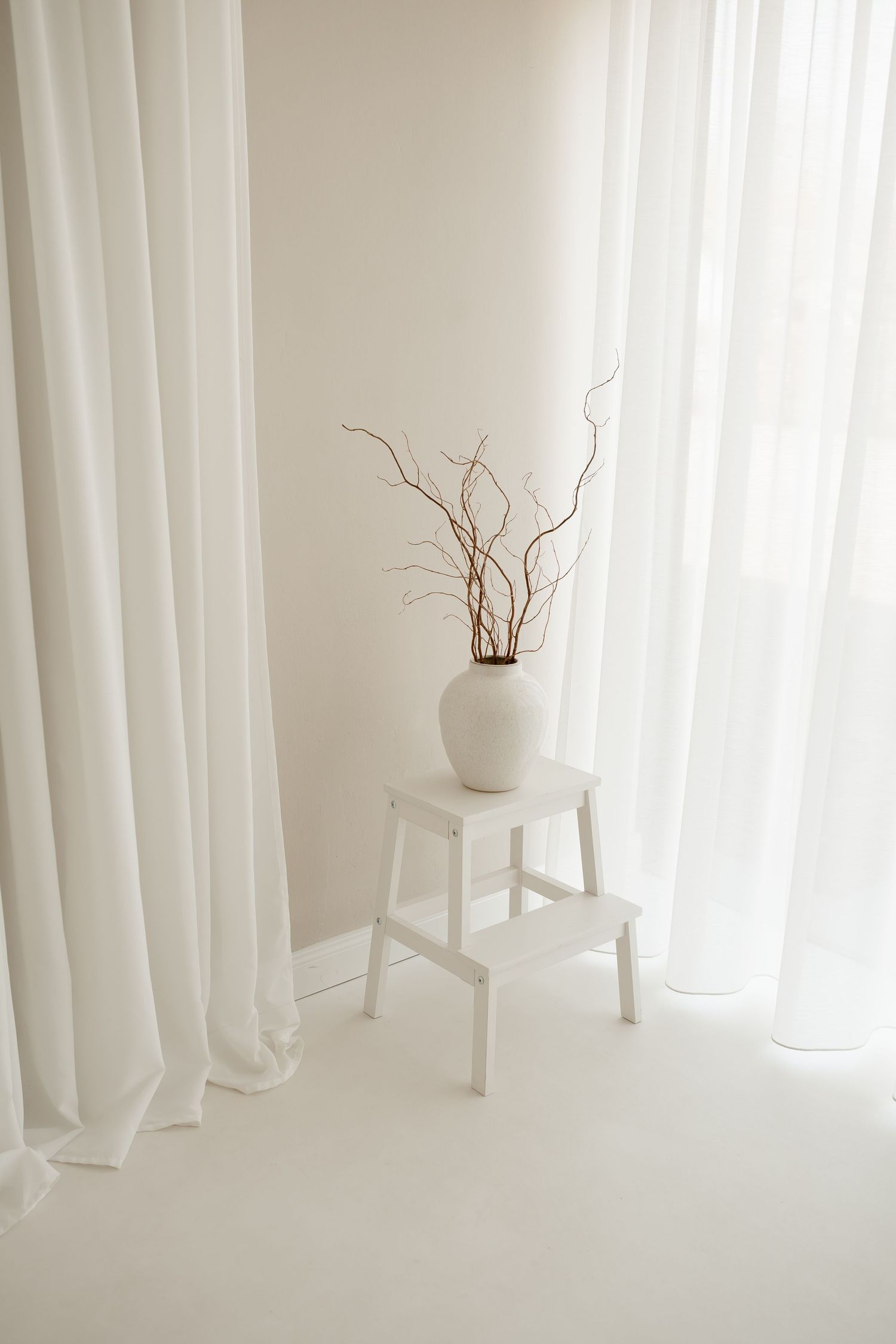 Minimalist white interior with dried branches in white vase on white stool near flowing sheer curtains.