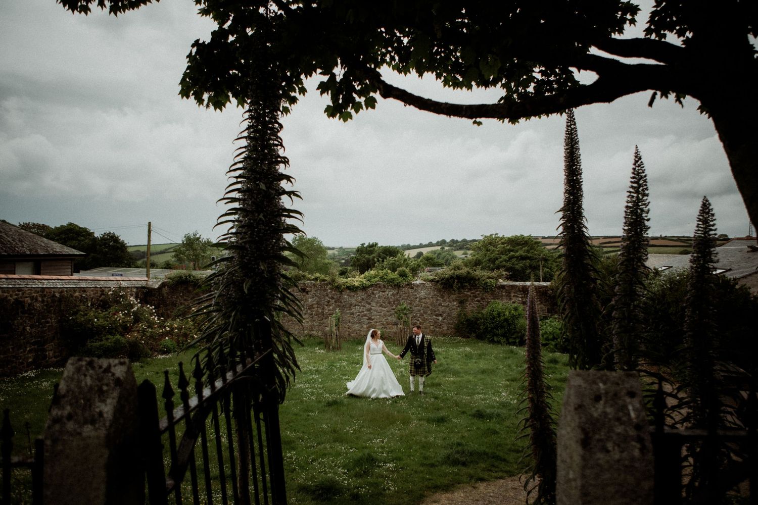 A romantic wedding portrait framed by tall palm trees on a lush green lawn against a moody sky.