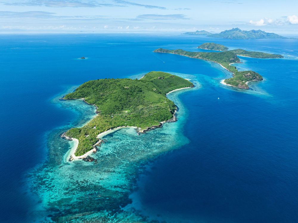 Aerial view of a tropical island surrounded by turquoise coral reefs and deep blue waters in the Pacific Ocean.