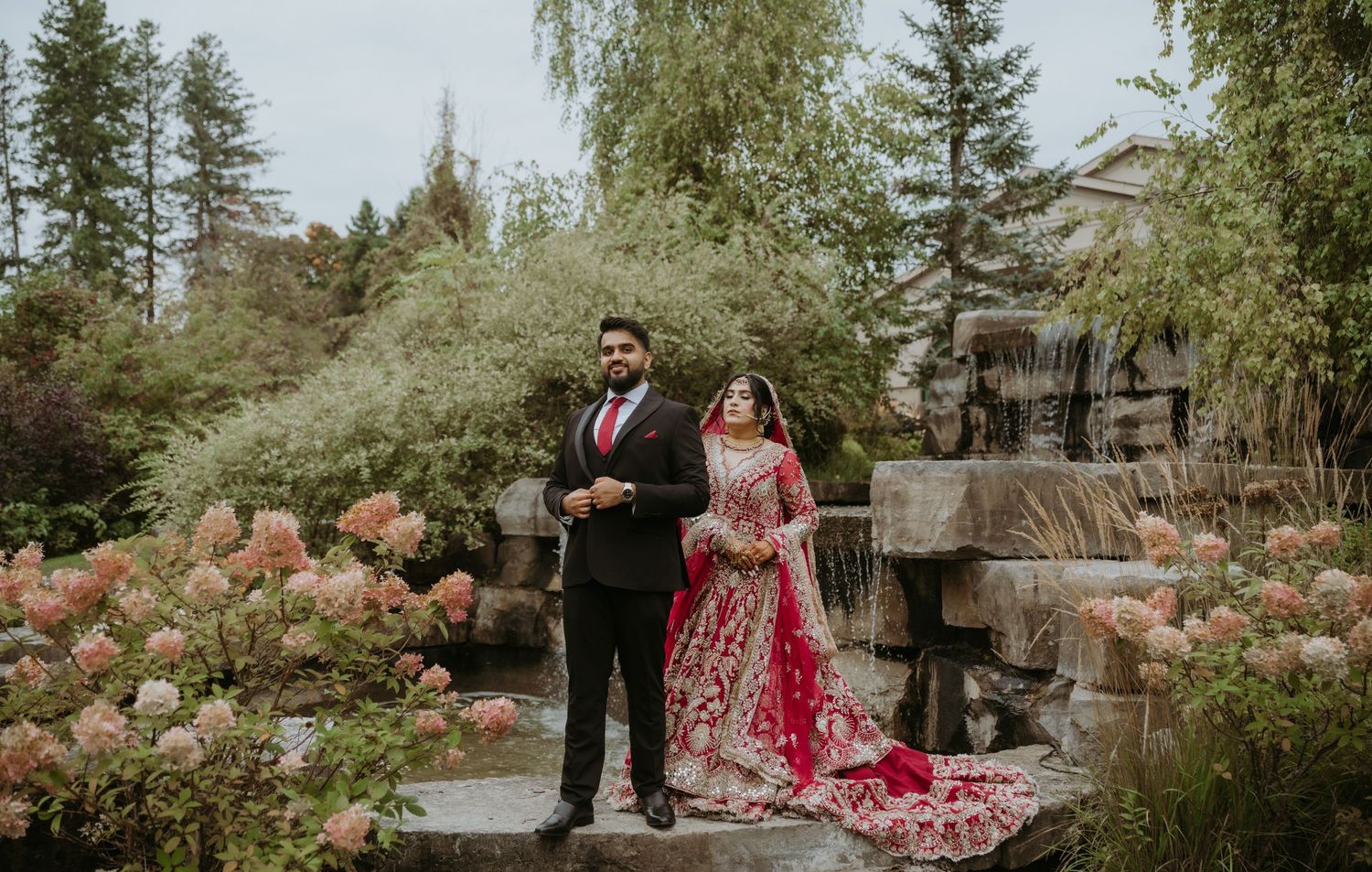 Couple in formal Indian attire pose in garden setting with stone waterfall and pink flowers.