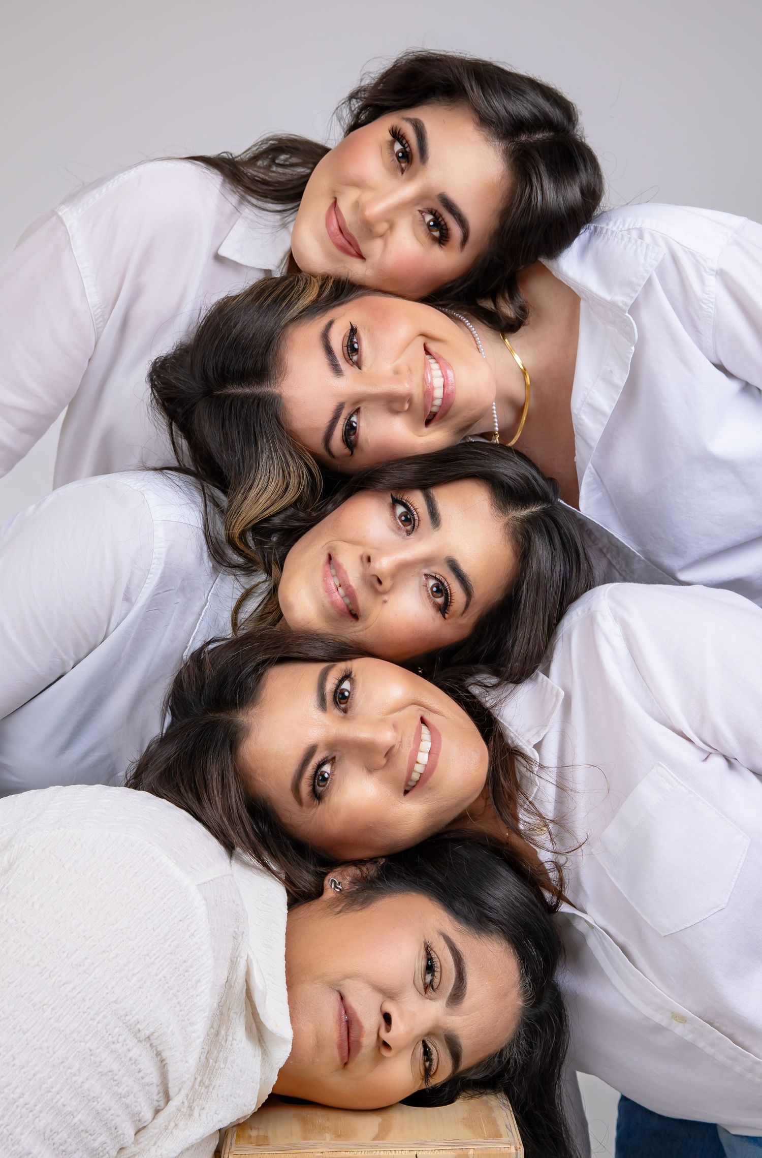Five people wearing white tops lying down with heads together in a circle formation while smiling at camera.
