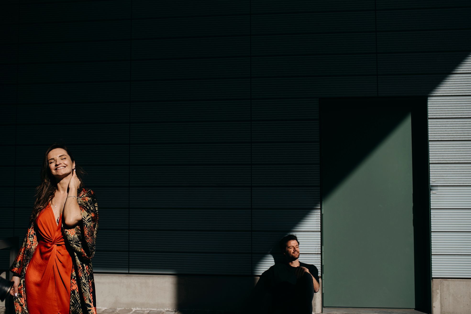 Woman in red kimono poses in dramatic sunlight while man stands in shadows behind her.