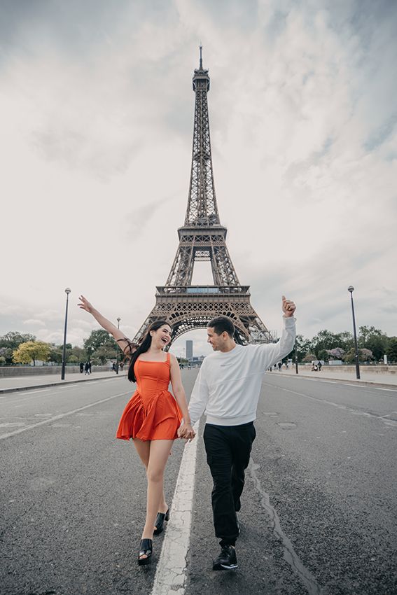 Joyful couple in red dress and white hoodie walking hand-in-hand before the Eiffel Tower.