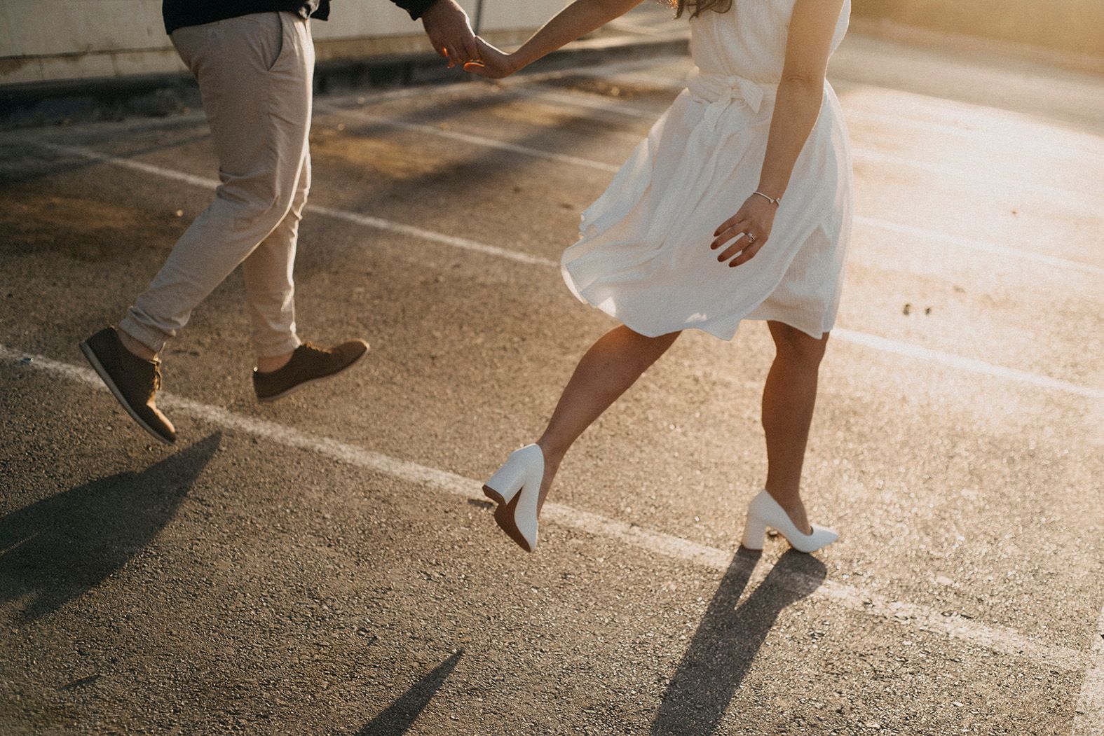 Couple holding hands and walking together in sunlight, bride wearing white dress and heels.