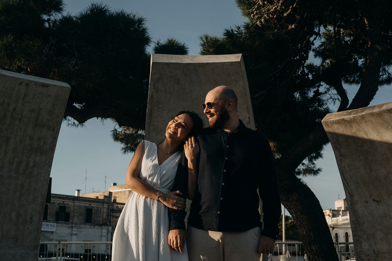 Smiling couple embracing outdoors near stone sculptures with trees and cityscape in background.