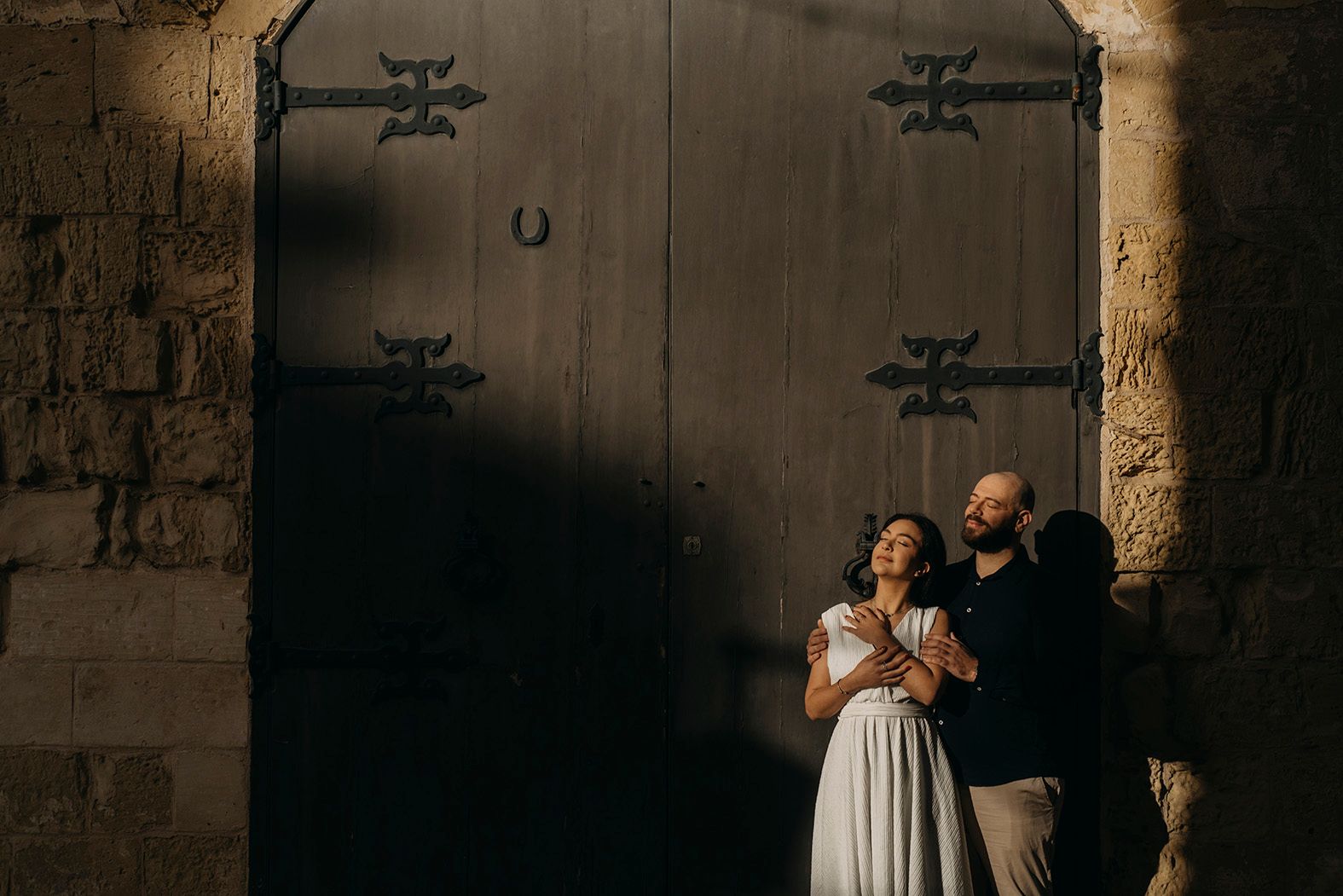 Romantic couple embracing before a large ornate wooden door in warm golden light.