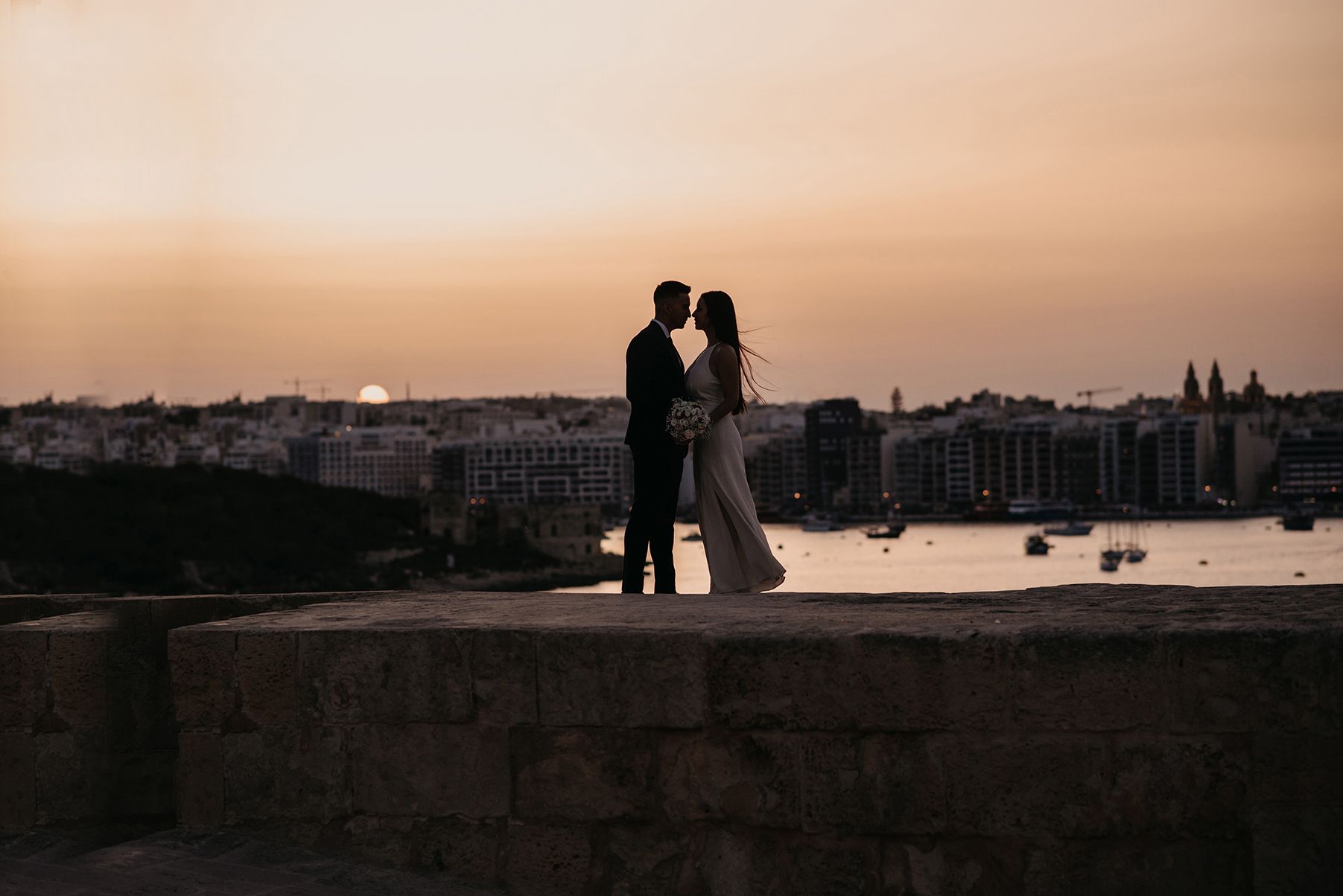 Silhouette of wedding couple kissing at sunset overlooking a beautiful coastal cityscape.