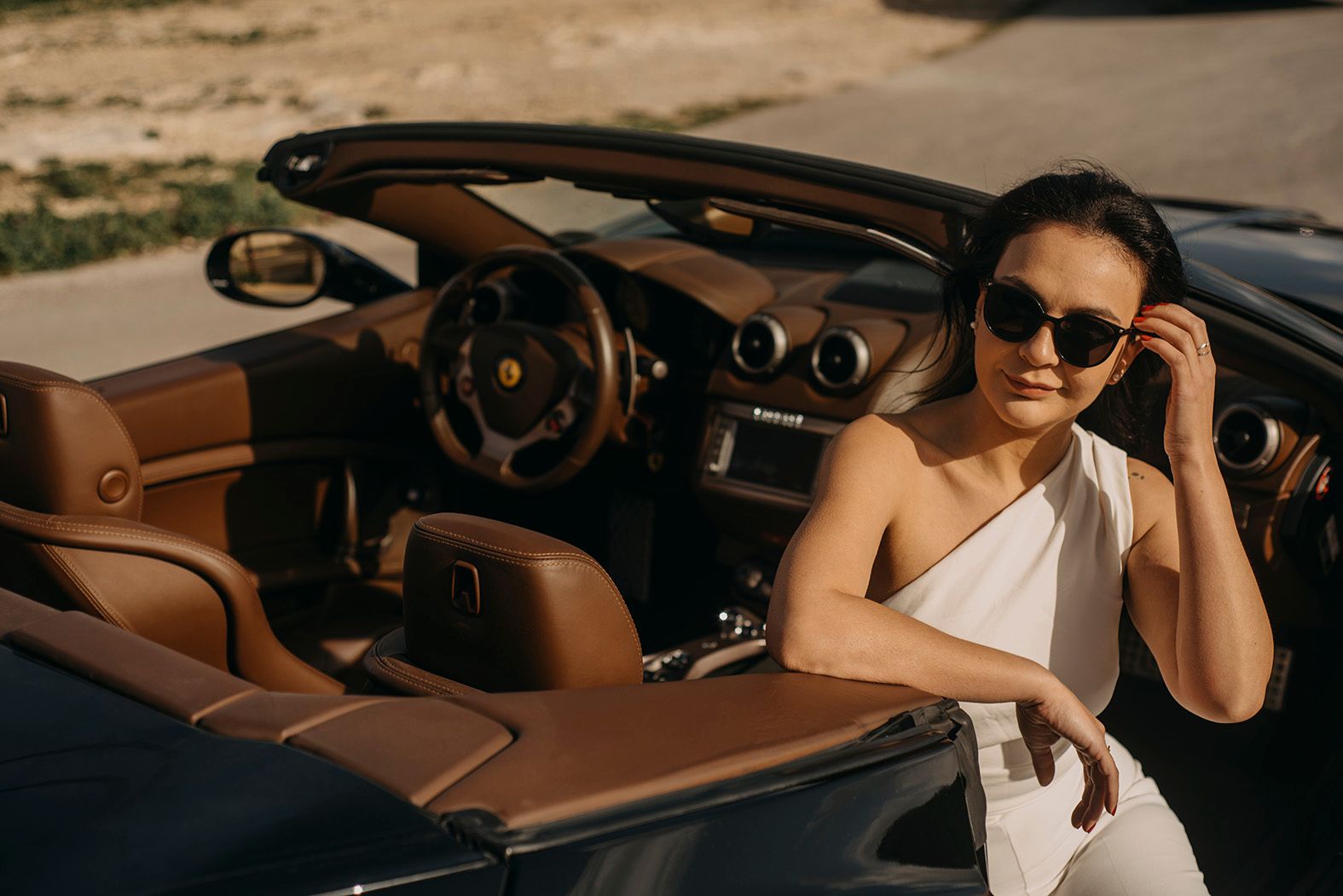 Stylish woman in white outfit wearing sunglasses sitting in a luxury Ferrari convertible.