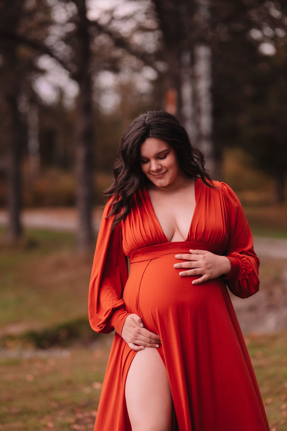 Expectant mother in flowing red dress poses in autumn forest setting.