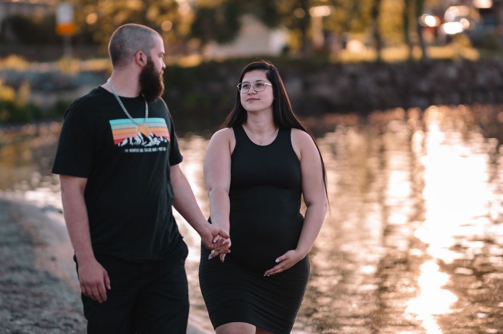 Expecting couple holding hands by water at sunset in casual attire.