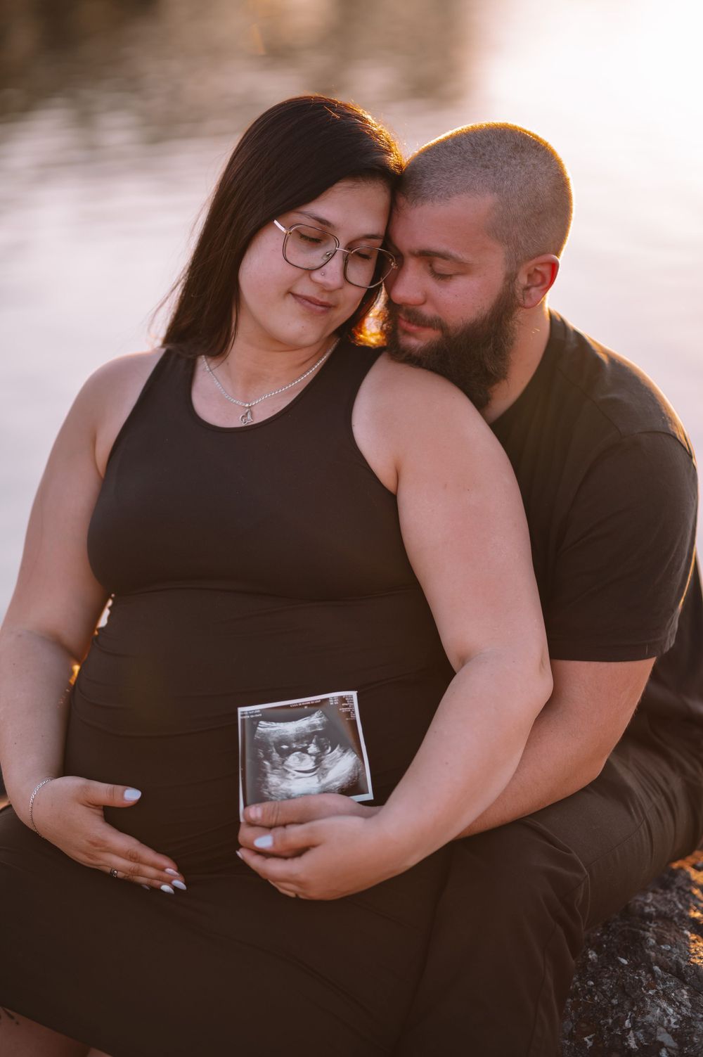 Expecting couple holding ultrasound photo during intimate waterfront photoshoot.