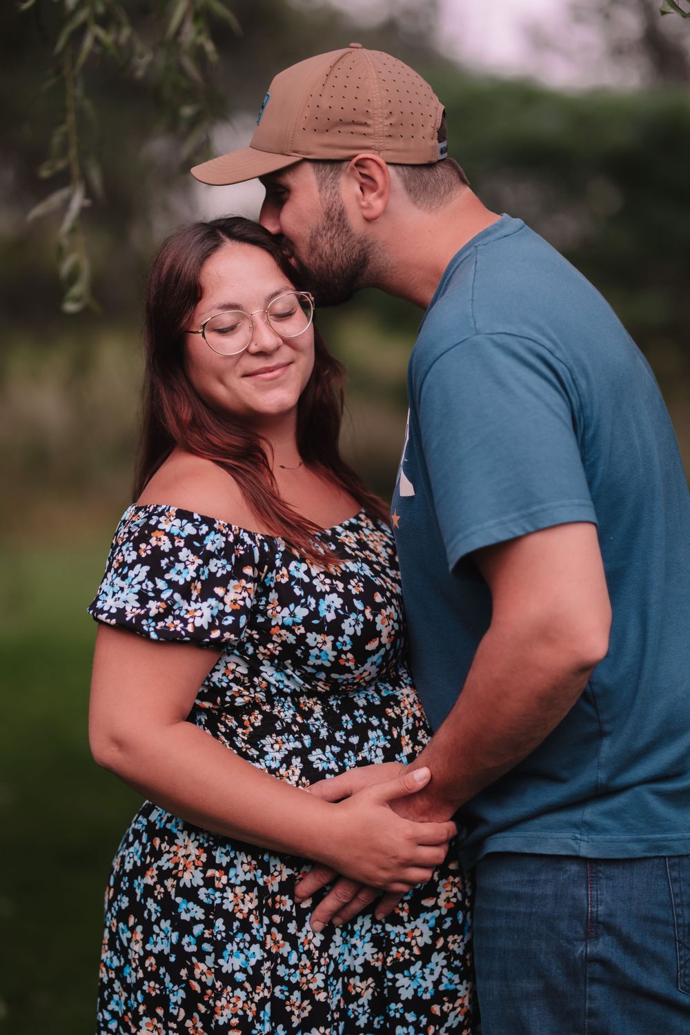 Intimate maternity portrait of couple in floral dress outdoors under willow tree.