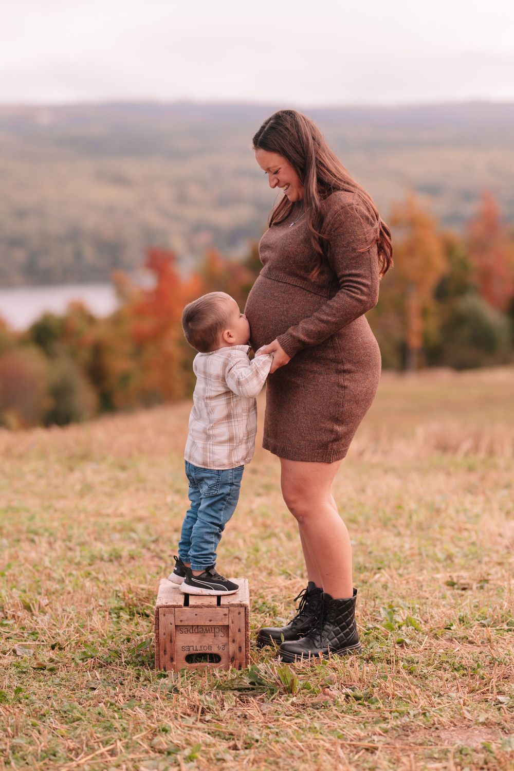 Child stands on wooden crate beside expectant mother in outdoor setting with lake view.