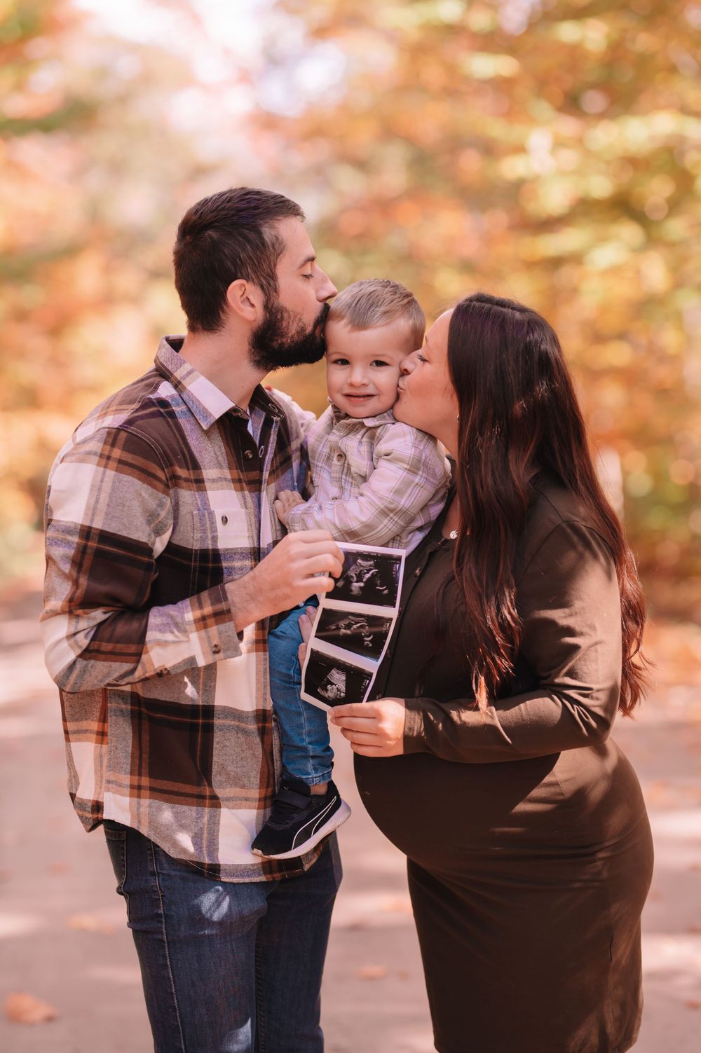 Family shares tender moment while holding ultrasound photos during autumn photoshoot.