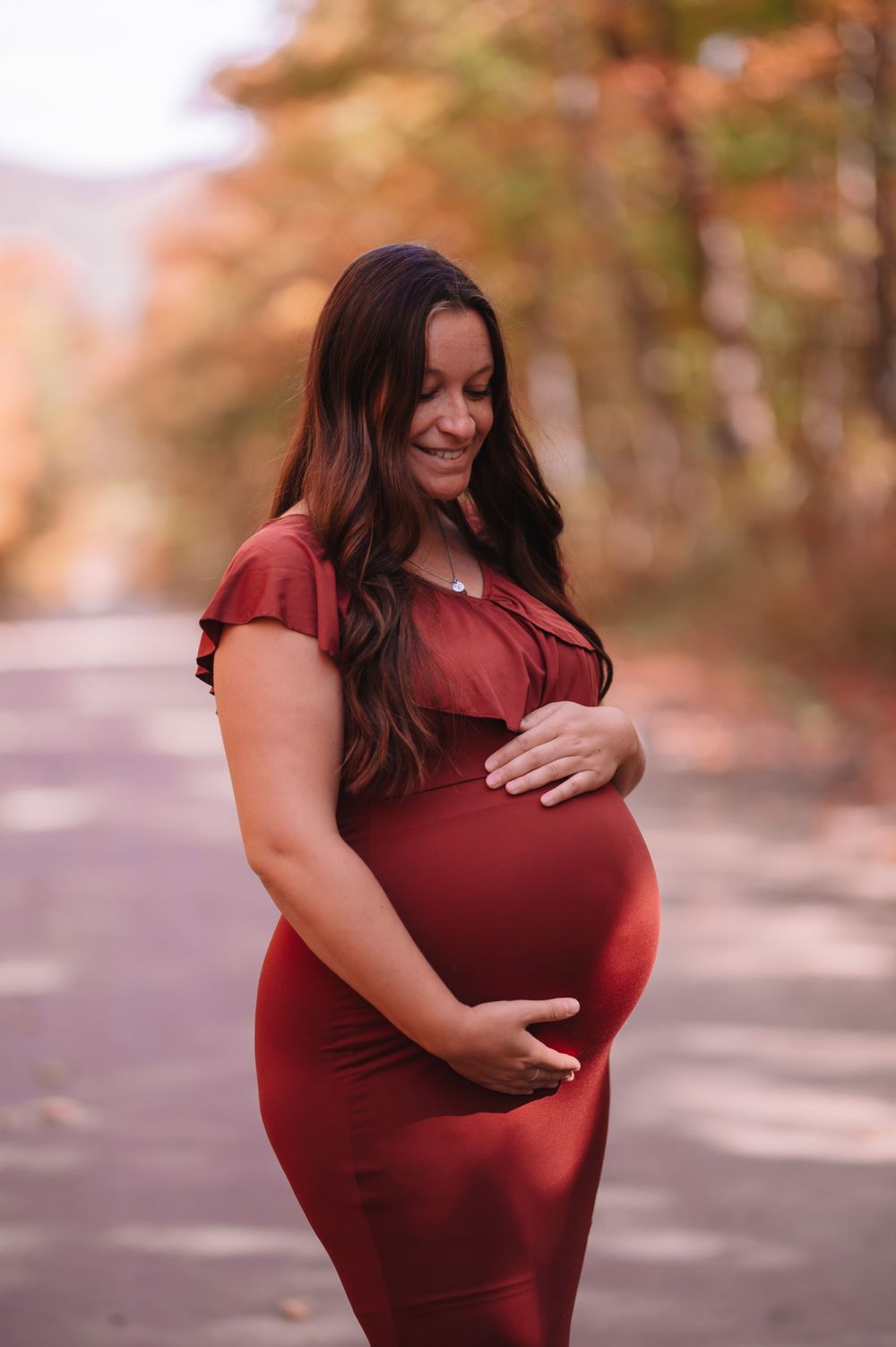 Expectant mother in burgundy dress poses on autumn path during maternity photoshoot.