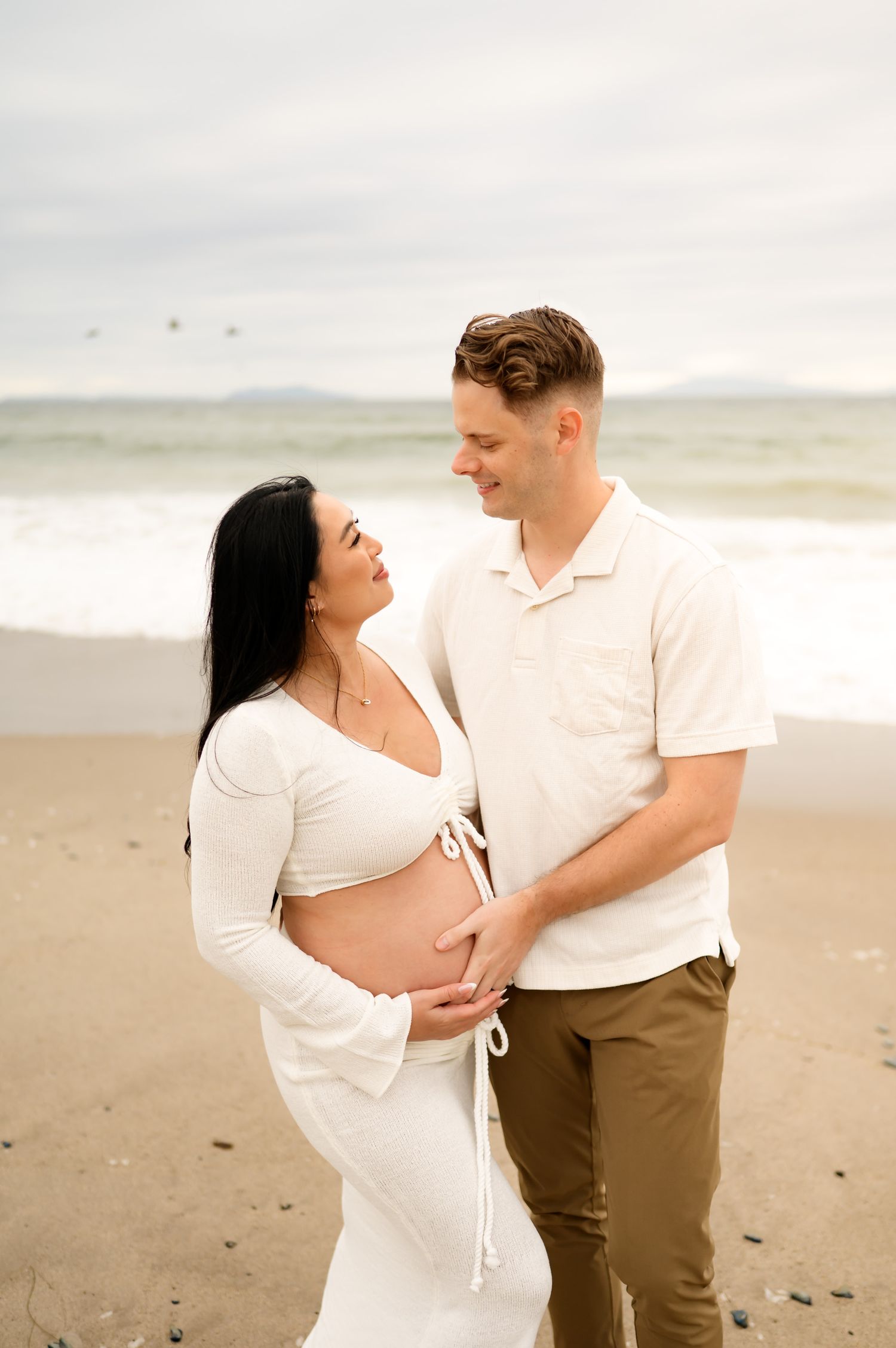couple stands by ocean on Ventura Beach during maternity photoshoot holding wife's bare belly while on their Santa Barbara babymoon