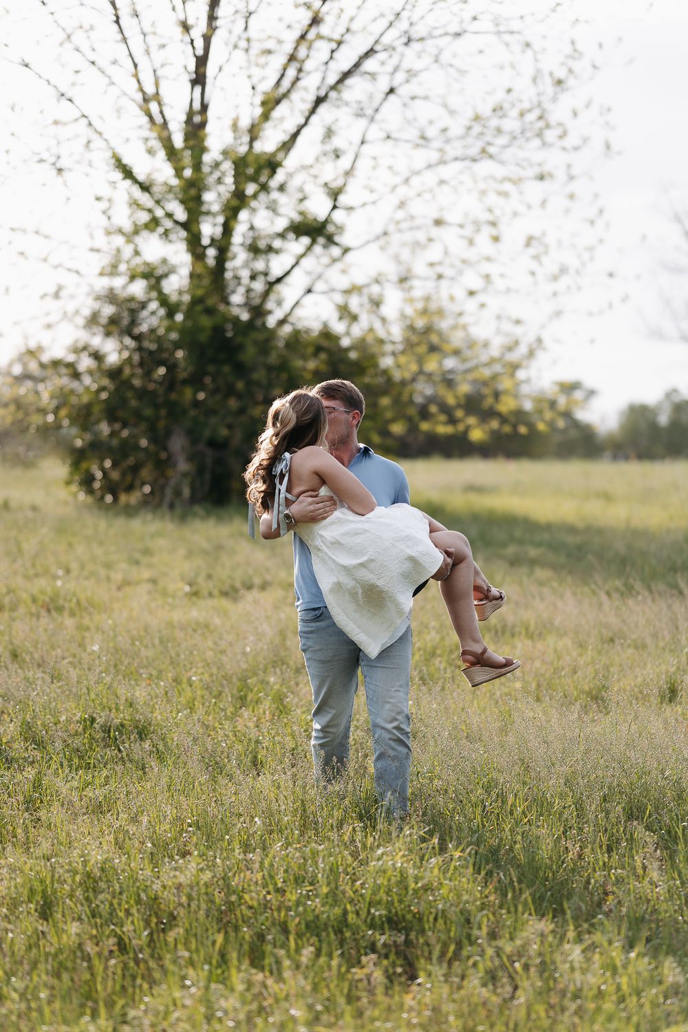 A man lifts a woman in a white dress in a green field, sharing a romantic moment near trees.