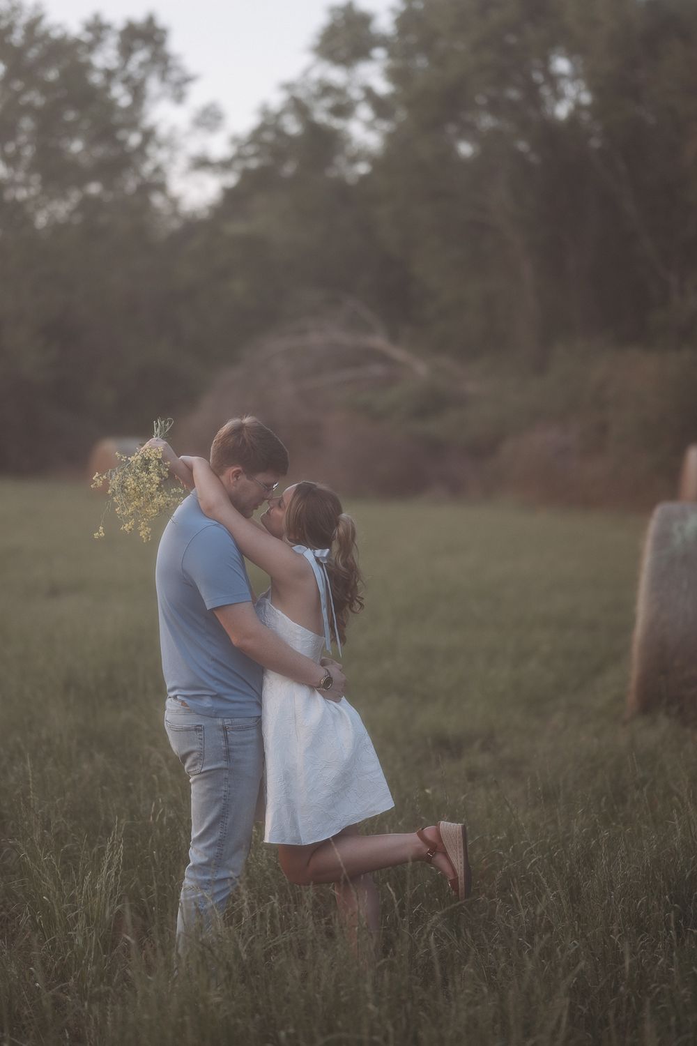 A couple embraces romantically in a green field at dusk, the woman holding wildflowers while lifting one leg.