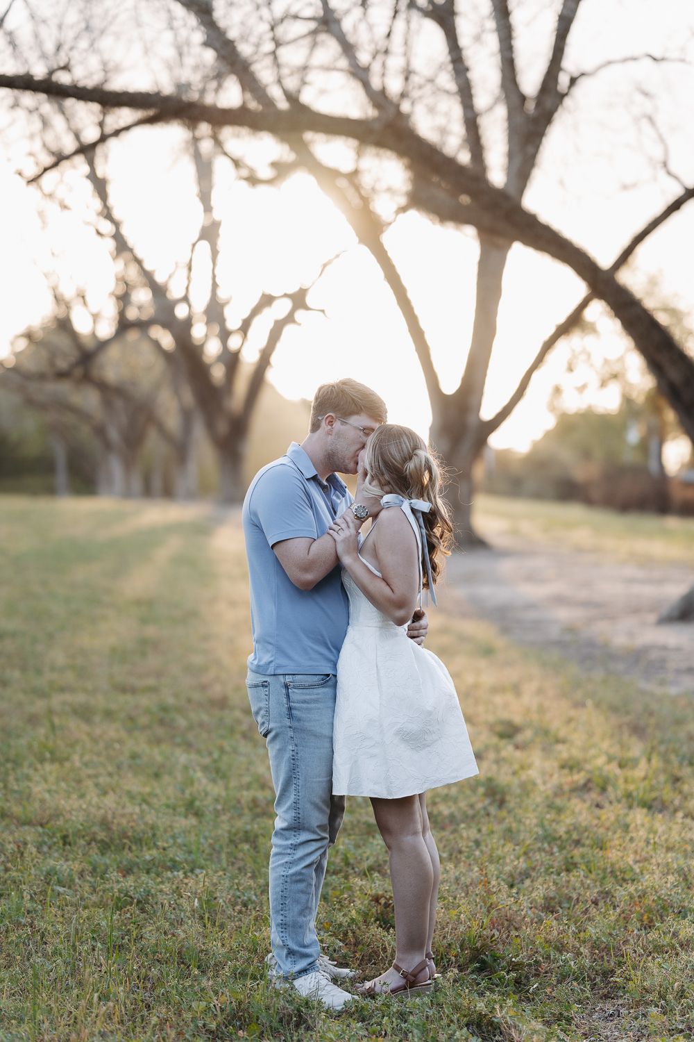 Couple sharing a romantic kiss at golden hour in a park with bare trees and warm sunlight glowing behind them.