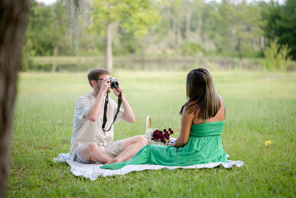 A couple shares a picnic on a blanket while taking photos in a grassy field.
