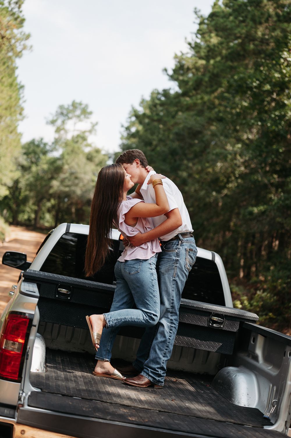 A couple shares an intimate moment sitting on the tailgate of a black pickup truck in a wooded setting.