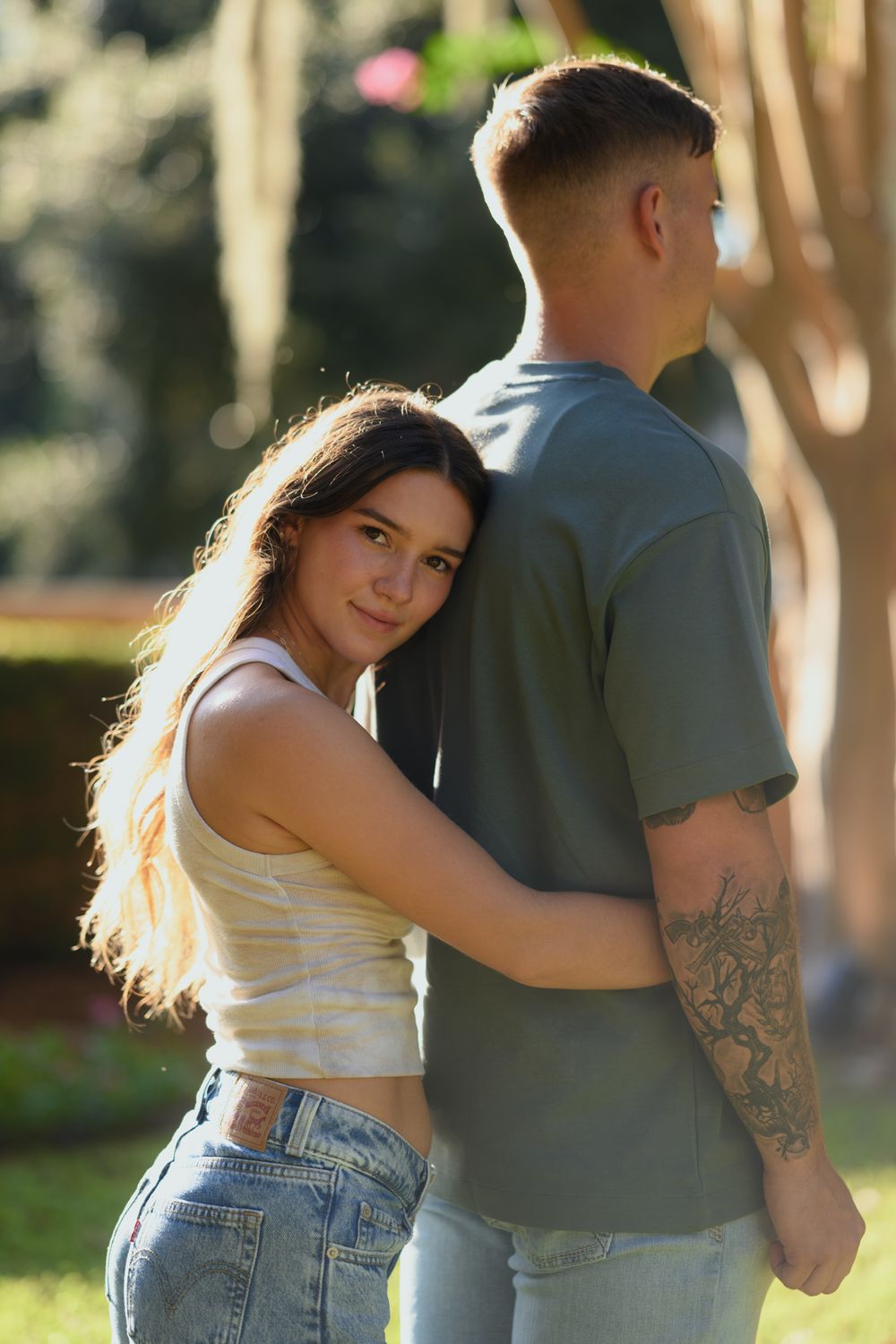 Couple in casual outfits embracing outdoors with sunlit trees in background.