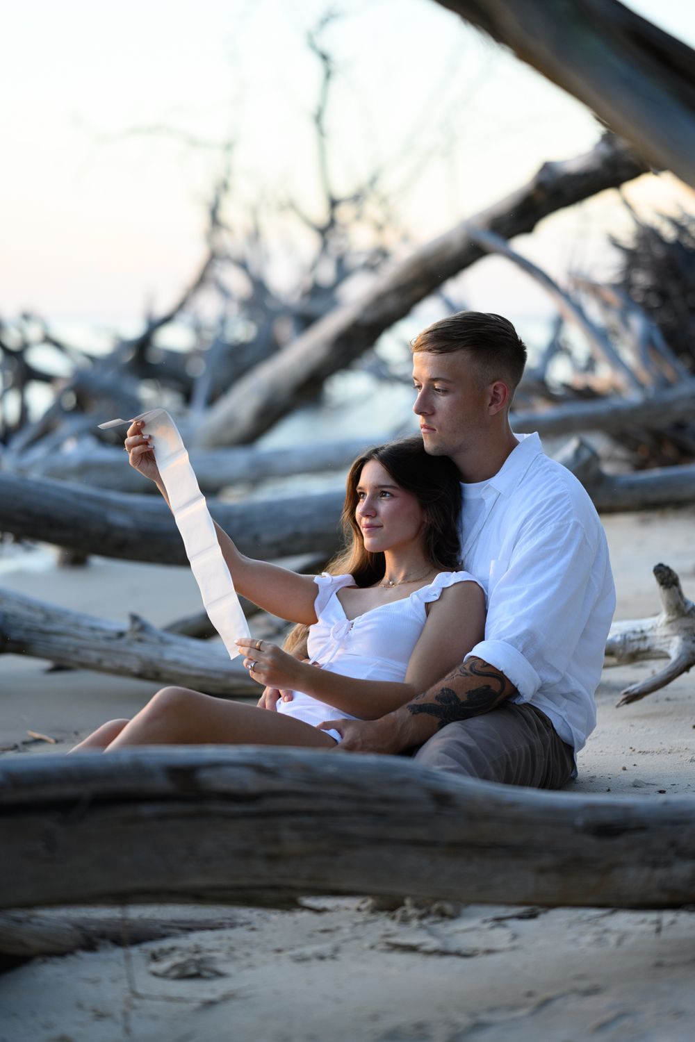 A couple sits together on driftwood at the beach during sunset reading a document.