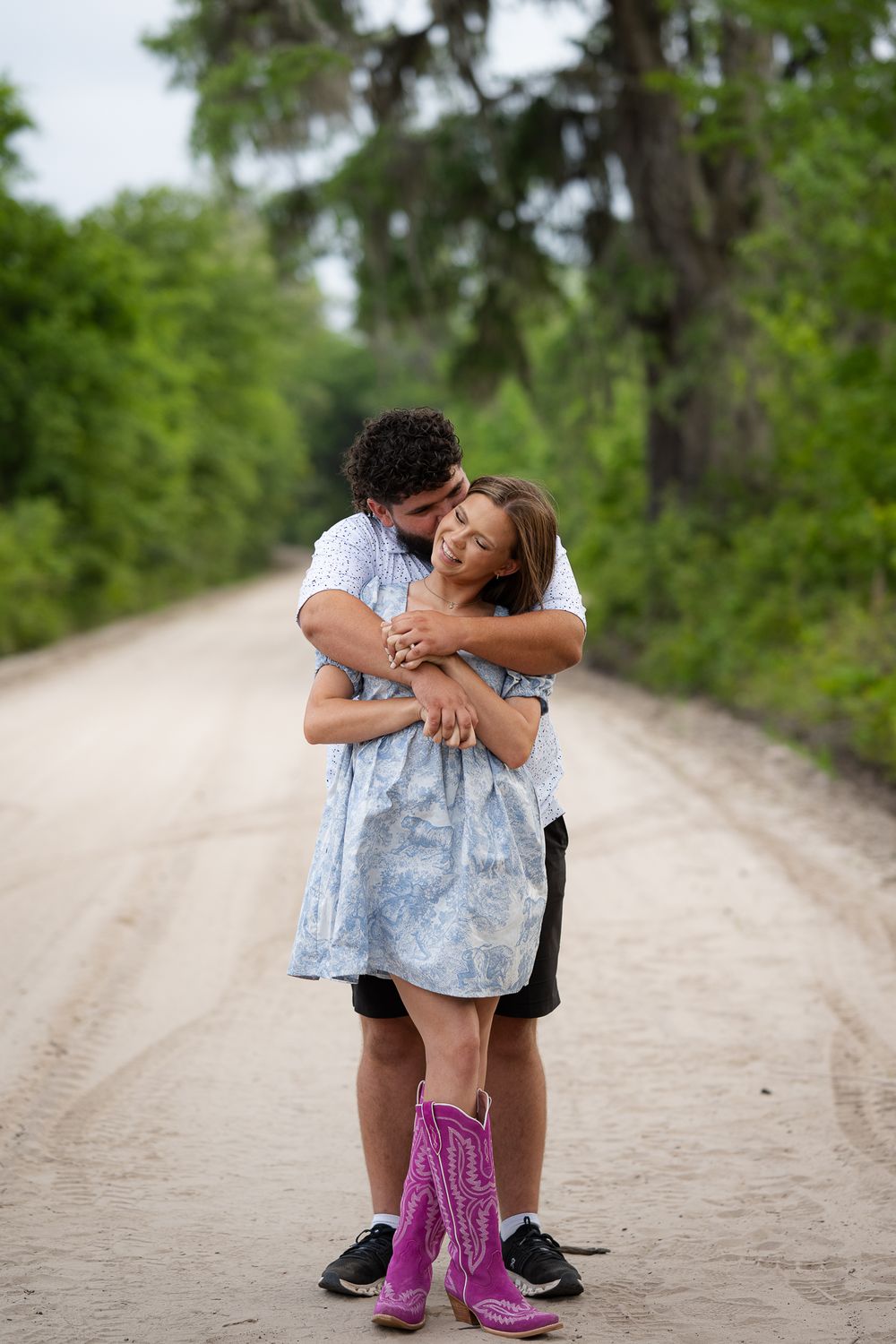 Two people share an intimate moment together on a sandy path surrounded by lush green trees and foliage.