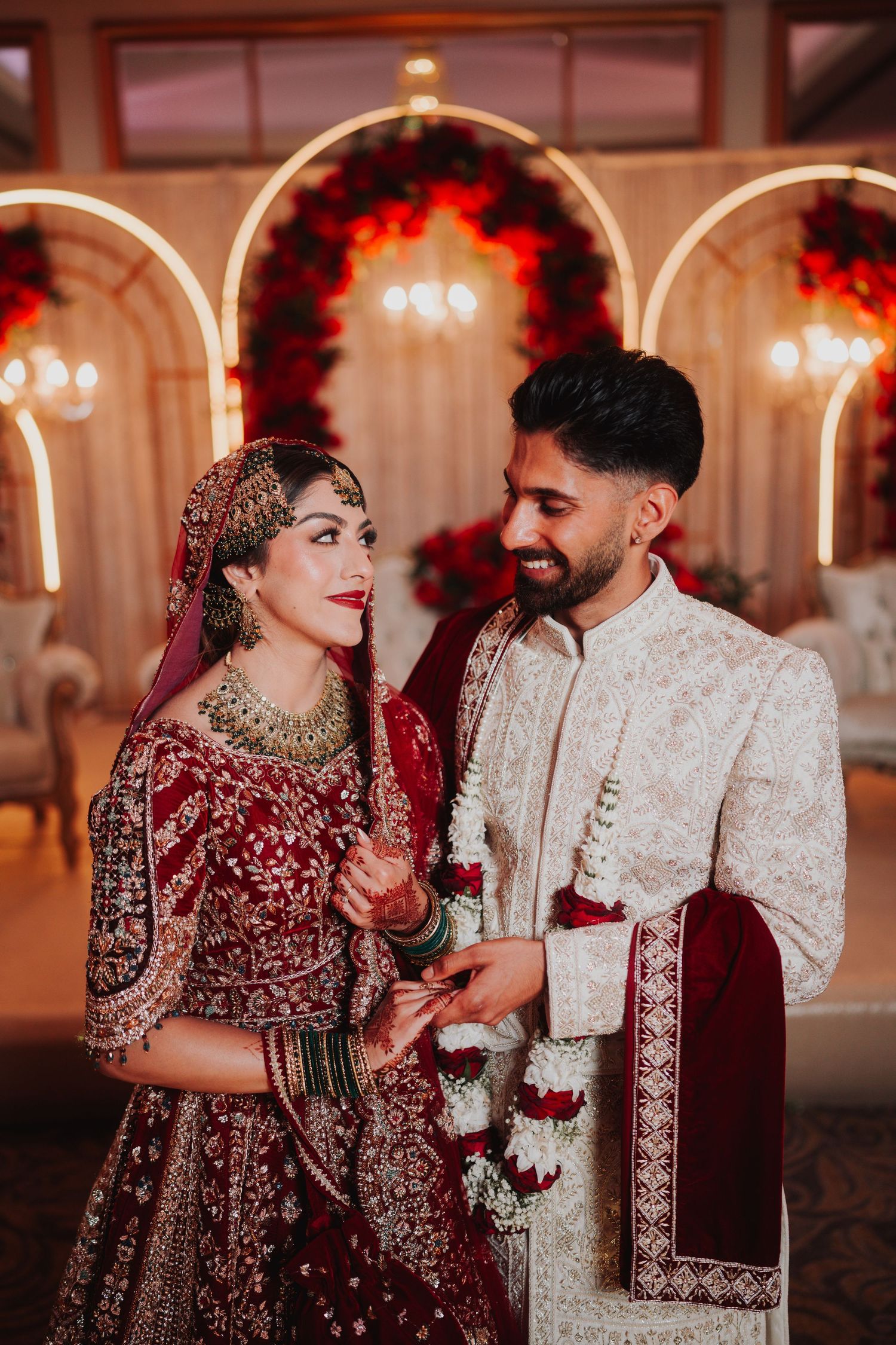 Traditional South Asian wedding couple in ornate red and cream outfits sharing a moment under lit arches.