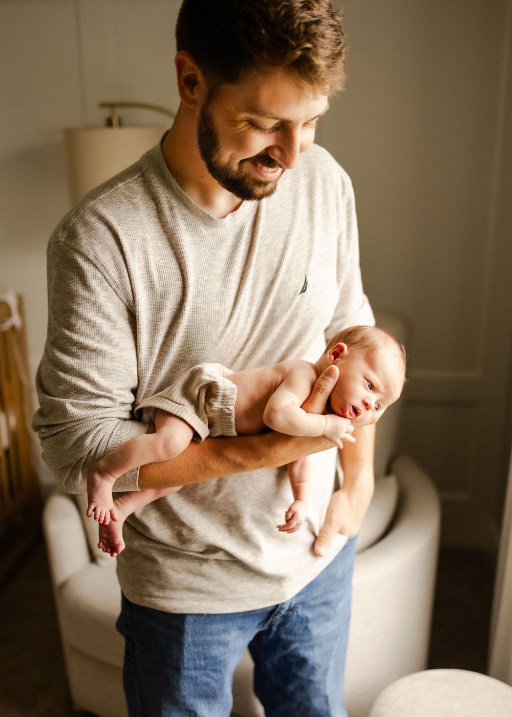 A person in a grey sweater smiles while holding a newborn baby in a warmly lit room.